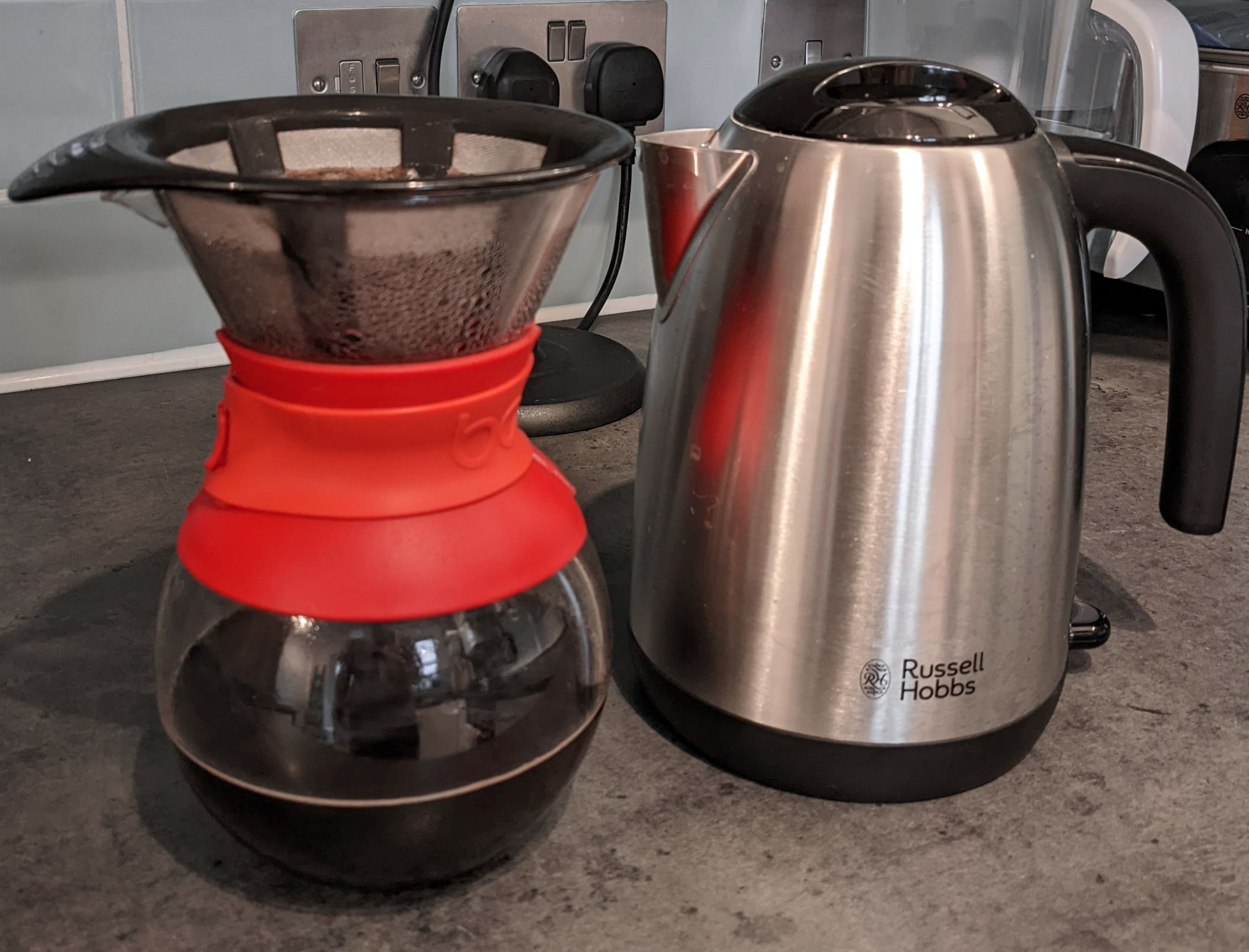 A Bodum pour over coffee maker with a red band next to a Russell Hobbs stainless steel kettle.