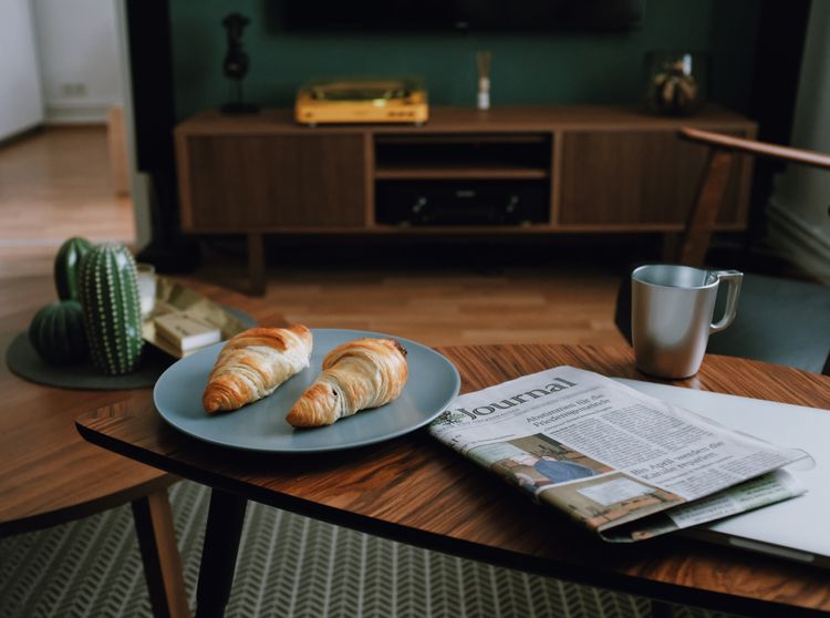 Croissants on a plate next to a newspaper and cup of coffee in a trendy living room.