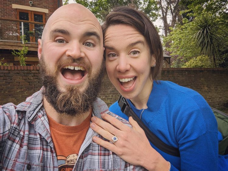 A bearded man in a grey checked shirt with his new fiance, both are smiling. 
